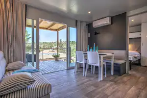 Bungalow-Terrasse mit Holzboden, zwei weißen Stühlen und Tisch, Blick durch große Fenster auf die Umgebung.