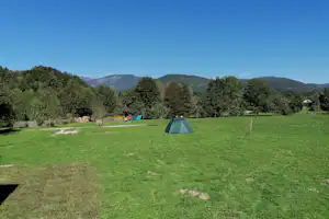 Zelt auf einer grünen Campingwiese, im Hintergrund Bäume, Berge und ein klarer Himmel.