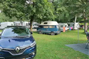 Blauer Renault vor einem Stellplatz, daneben ein Wohnmobil und geparkte Autos auf grünem Rasen zwischen Bäumen.