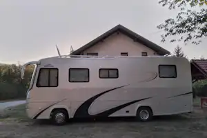 Weißes Wohnmobil steht auf dem Campingplatz vor einem Haus mit Giebel, im Hintergrund Bäume und ein Weg.
