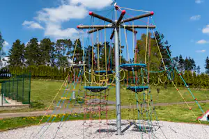 Spielplatz mit Seilnetz-Schaukeln und bunten Kletterseilen an einer Stahlstange, Rasen und Bäume im Hintergrund.