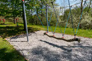 Seilnetz-Spielgerät auf einem Campingplatz mit Kiesboden, Pfosten und umgebenden Bäumen im Hintergrund.