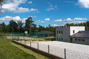 Moderne Campinganlage mit weißen Gebäuden, gepflastertem Weg, umzäuntem Sportplatz und Rasenfläche mit Bäumen im Hintergrund.