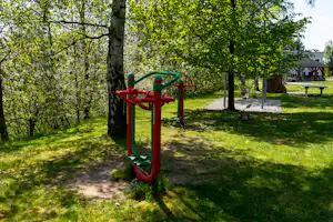 Rotes Spielgerät auf einem Kiesweg im grünen Campingbereich, umgeben von Bäumen und einer Bank mit Tisch.