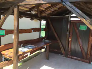 Holzunterstand mit Bank und Tisch, seitlich ein Grillbereich, dahinter eine offene Tür und grünes Gelände.