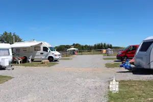 Wohnwagen und Campingfahrzeuge auf einem Stellplatz, Schotterfläche, Bäume und Zaun im Hintergrund.