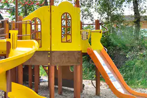 Gelbes Spielgerät mit Rutsche und Kletterelementen auf Holzpodest, im Garten mit Bäumen und Hecke.
