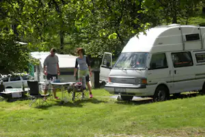Wohnmobil am Campingplatz, daneben Tisch und Stühle im Schatten der Bäume, zwei Personen am Grillbereich.