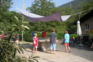 Terrasse am Campingplatz mit Sonnensegel, Kindern am Tisch und Bäumen im Hintergrund, daneben ein Grill und Stühle.