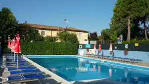 Schwimmbad mit blauem Wasser, Liegen und Sonnenschirmen vor einem Campinggebäude, Hecke und Bäume im Hintergrund.