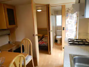 Innenansicht einer Ferienwohnung im Campingbereich mit Küchenzeile, Esstisch und Stühlen sowie Blick in das Bad mit Toilette.