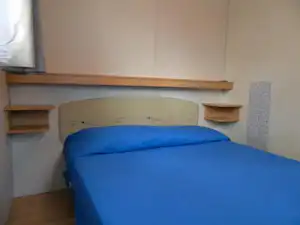 Bett mit blauem Bettbezug in einem einfachen Campingzimmer, mit Holzregalen an der Wand und grauem Wandpaneel.