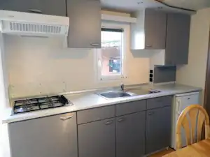 Kleine Küchenzeile mit Herd, Spüle und Schränken, Fenster mit Blick nach draußen, Holzstuhl am Tisch.