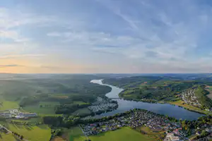 Blick auf ein Flusstal mit Campingplatz am Ufer, Wohnhäusern und Feldern in der grünen Landschaft.