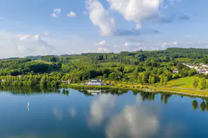 Ruhiger See mit Spiegelung, bewaldete Hügel und Häuser am Ufer, Blick von oben.