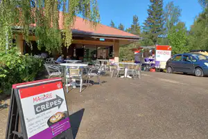 Terrasse am Camping-Restaurant mit Tischen und Stühlen, Verkaufswagen und Auto vor dem Gebäude, umgeben von Bäumen.
