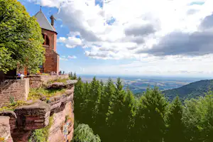Steinbau mit Turm und Balkon, umgeben von Bäumen, Blick über das weite Tal unter wolkigem Himmel.