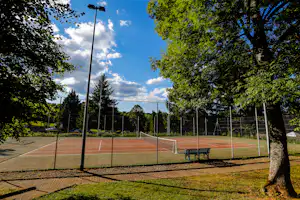 Tennisplatz auf dem Campinggelände mit Umzäunung, Bank und Bäumen am Rand, daneben ein Weg und Laternen.
