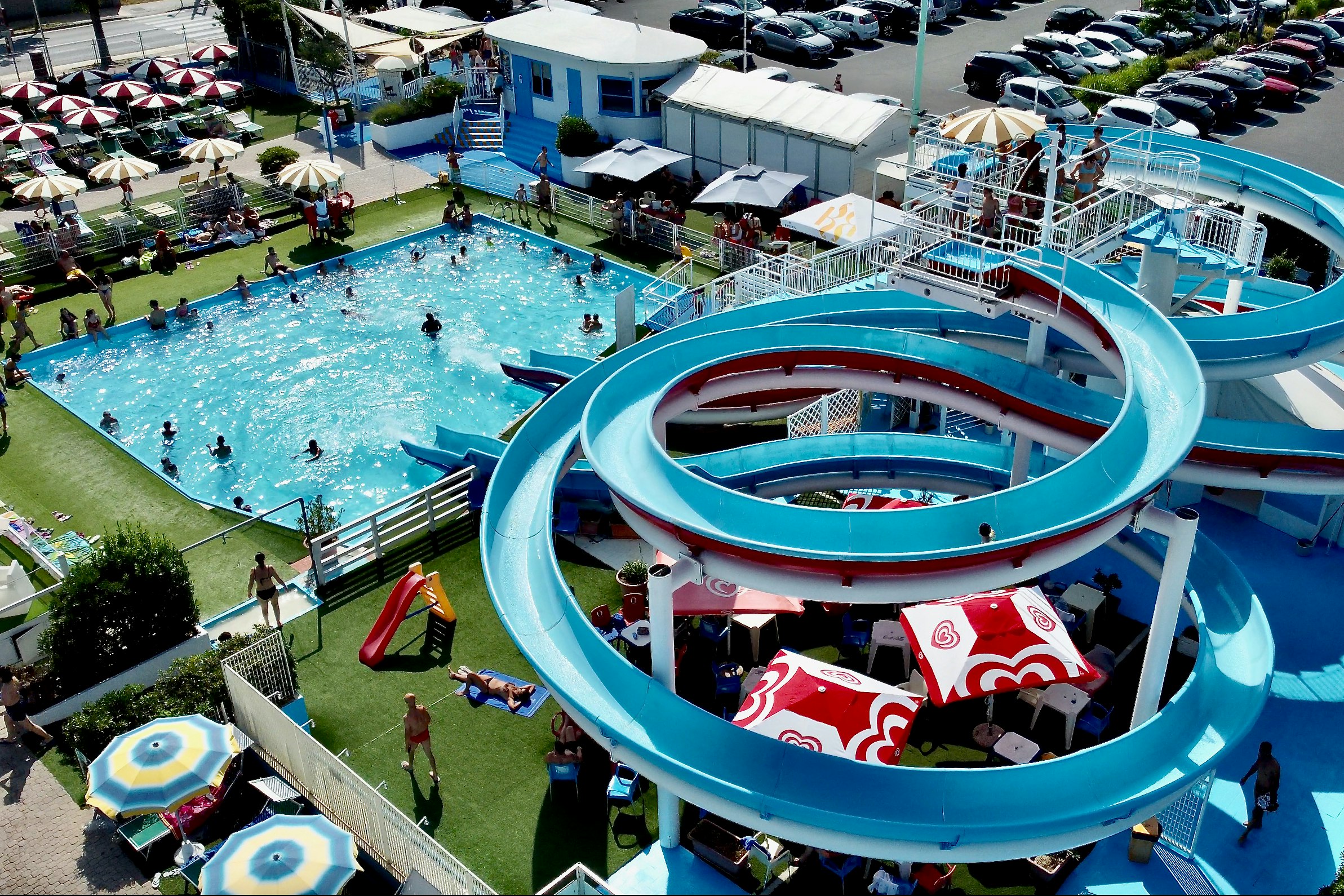 Großer Wasserpark mit mehreren blauen Rutschen, Kinderbecken und Liegen auf dem Campingplatz, daneben Wege und Zäune.