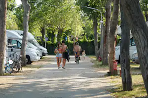 Campingplatz-Gasse im Schatten der Bäume, Wohnmobile und ein Fahrrad, mehrere Personen gehen am Weg entlang.
