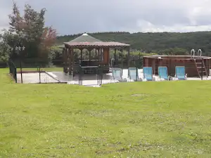 Überdachte Terrasse am Campingplatz mit Pool, mehreren Liegestühlen und Zaun, im Hintergrund Bäume und bewaldete Hügel.