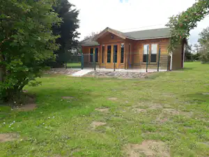 Holzhaus auf dem Campingplatz mit großen Glasfronten, umzäuntem Bereich und Rasenfläche, umgeben von Bäumen.