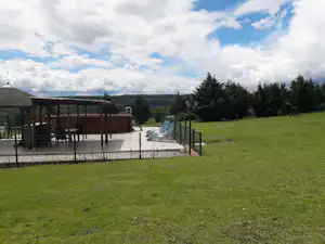 Terrasse am Campingplatz mit blauen Liegestühlen, Holzüberdachung, umzäuntem Bereich und grünem Rasen vor weitem Tal.