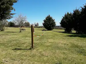 Grünes Gras auf einem Campingplatz mit Holzpfosten, Absperrband und zwei Picknickbänken im Hintergrund.
