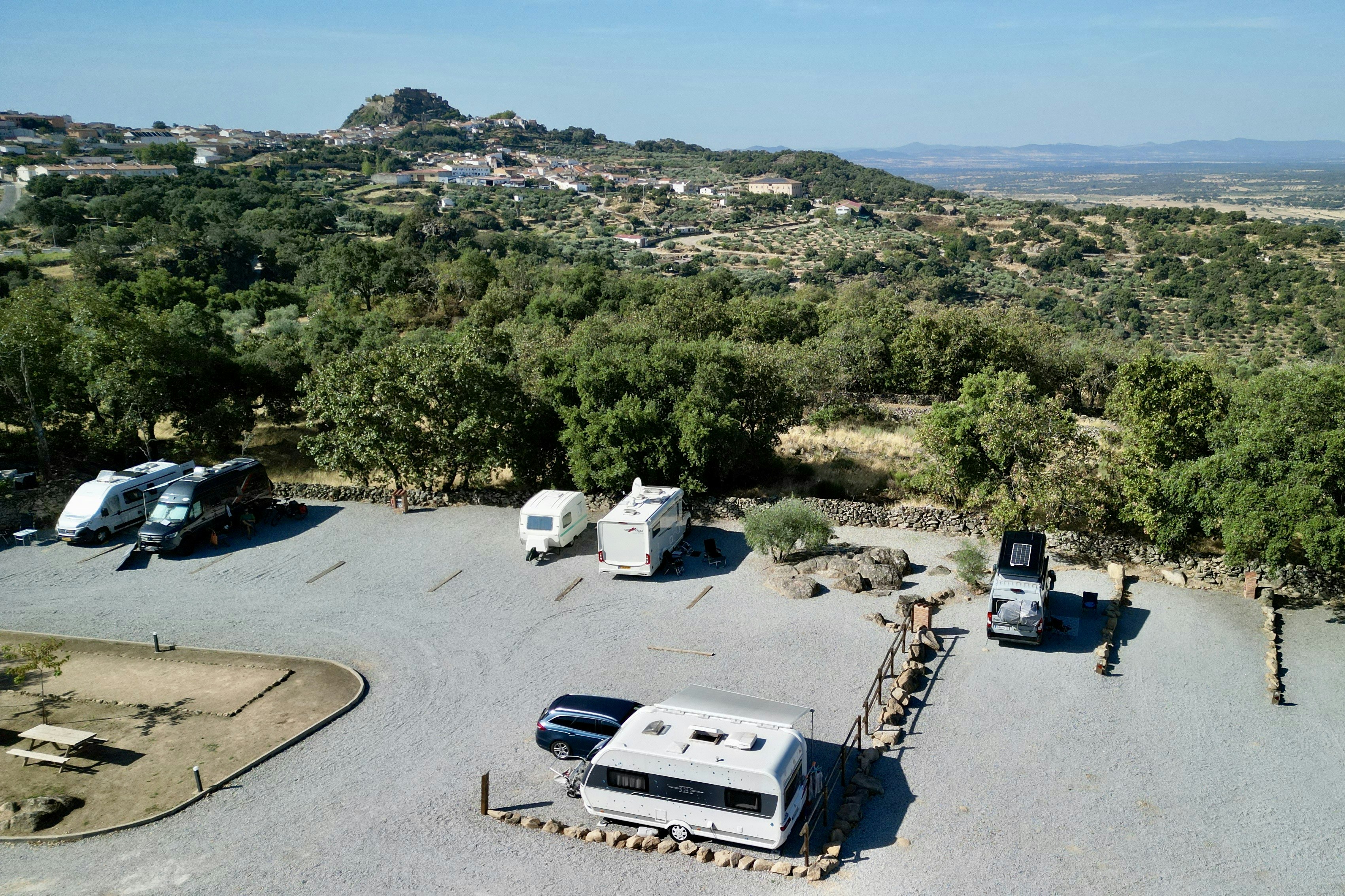 Wohnmobile und Campingfahrzeuge auf einem Stellplatz mit Betonfläche, umgeben von Bäumen und Hügeln, Blick ins Tal.