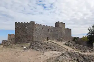 Eine alte Burganlage aus Stein mit Türmen und Mauern auf einem Hügel, umgeben von Felsen und einem Weg.