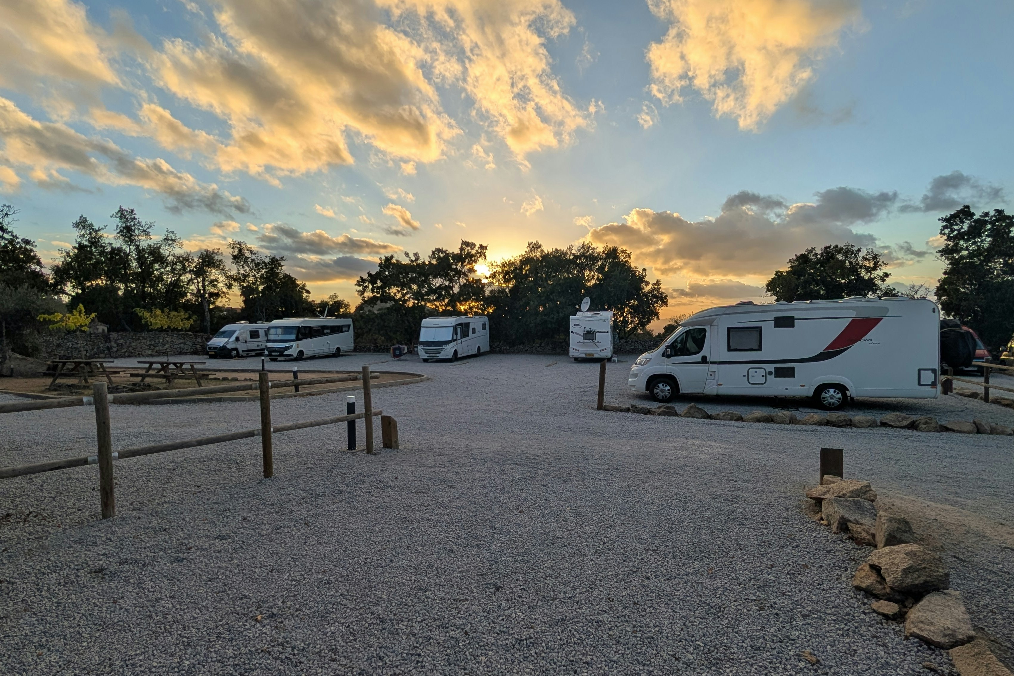 Wohnmobile auf einem Campingplatz, Kiesweg und Holzgeländer, im Hintergrund Bäume und ein Wohnwagen bei Sonnenuntergang.