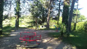 Roter Karussell-Spielplatz auf Kiesfläche, im Hintergrund Schaukelgestell und Bäume im Campinggelände.