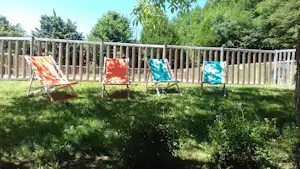 Drei Sonnenliegen im Campinggarten vor einem Holzlattenzaun, umgeben von grünem Rasen und Büschen.