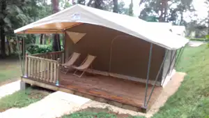 Campingzelt auf Holzplattform mit Veranda und zwei Liegestühlen, umgeben von Rasen und Bäumen im Hintergrund.