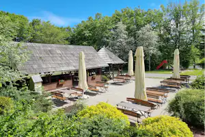 Terrasse mit Holzbänken und Sonnenschirmen vor mehreren Ferienhütten, umgeben von Bäumen und gepflegten Sträuchern.