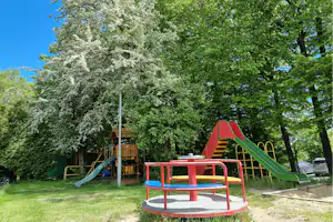 Spielplatz mit Karussell und Rutsche im Campingbereich, umgeben von Bäumen und Rasenfläche unter blauem Himmel.