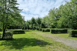 Grünes Campinggelände mit Rasenfläche, Kiesweg und Hecken, umgeben von Bäumen und Laternen im Hintergrund.