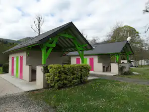 Campinggebäude mit überdachter Terrasse, grünen Holzstützen und bunten Türen, davor Hecke und Rasenfläche.
