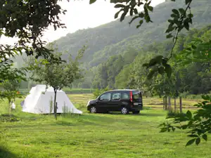 Zelt auf einer grünen Wiese mit geparktem Auto, umgeben von Bäumen und Blick auf bewaldete Hügel.