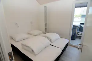 Schlafbereich mit Doppelbett, Kissen und Nachttischlampen; daneben ein offenes Regal und eine Balkontür zum Holzdeck.