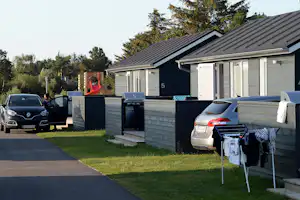 Campingunterkunft mit Terrasse, Grill und Autos direkt am Stellplatz, im Hintergrund Bäume und ein Schuppen mit Nummer 5.