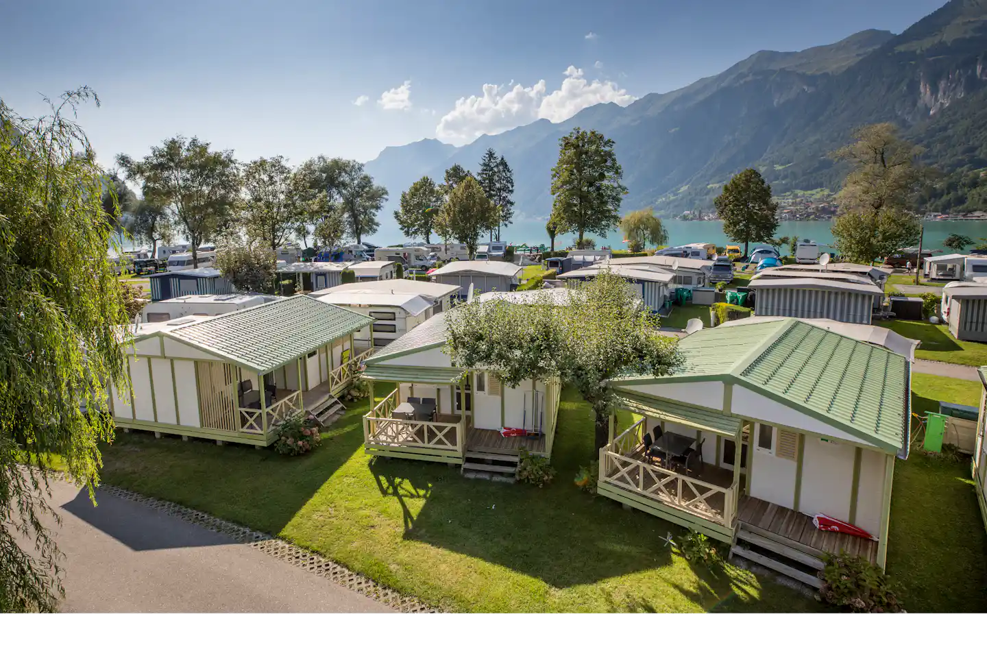 Reihe von Camping-Unterkünften mit grünen Rasenflächen, Holzterrassen und Zäunen, im Hintergrund Berge und ein türkisfarbenes Wasserbecken.