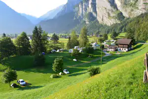 Campingplatz auf einer grünen Wiese mit Zelten, Autos und Wohnwagen, umgeben von Bäumen und Bergen im Tal.