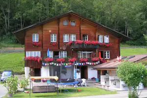 Holzhaus mit Terrasse, roten Blumenampeln, Wäscheleine und Gartenmöbeln vor Waldkulisse.