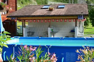 Blumen vor einem blauen Campingpool, dahinter ein Gebäude mit Dachfenstern und Terrasse im Garten.