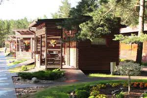 Holzunterkünfte und Sichtschutzwände am Campingplatz, mit gepflastertem Weg, gepflegtem Gartenbeet und Bäumen im Hintergrund.