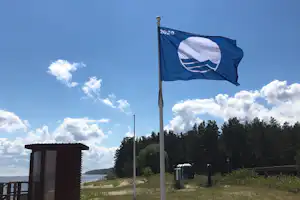 Campingplatz-Fahne auf einem Pfosten vor einem Weg, daneben ein Holzhaus und Rasen mit Bäumen im Hintergrund.
