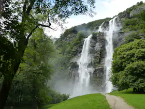 Großer Wasserfall im grünen Tal, umgeben von Bäumen und steilen Felswänden, mit Grasfläche und Weg im Vordergrund.