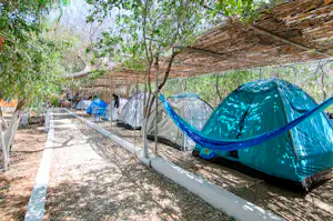 Blaues Zelt auf einem gepflasterten Stellplatz am Campingplatz, daneben weitere Zelte unter Schattendächern und Bäumen.