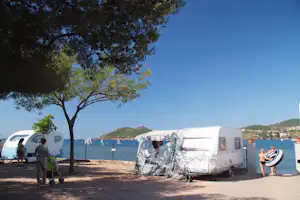 Zelte auf einem Campingplatz am Meer, im Hintergrund Wasser, Hügel und ein Baum am Ufer.